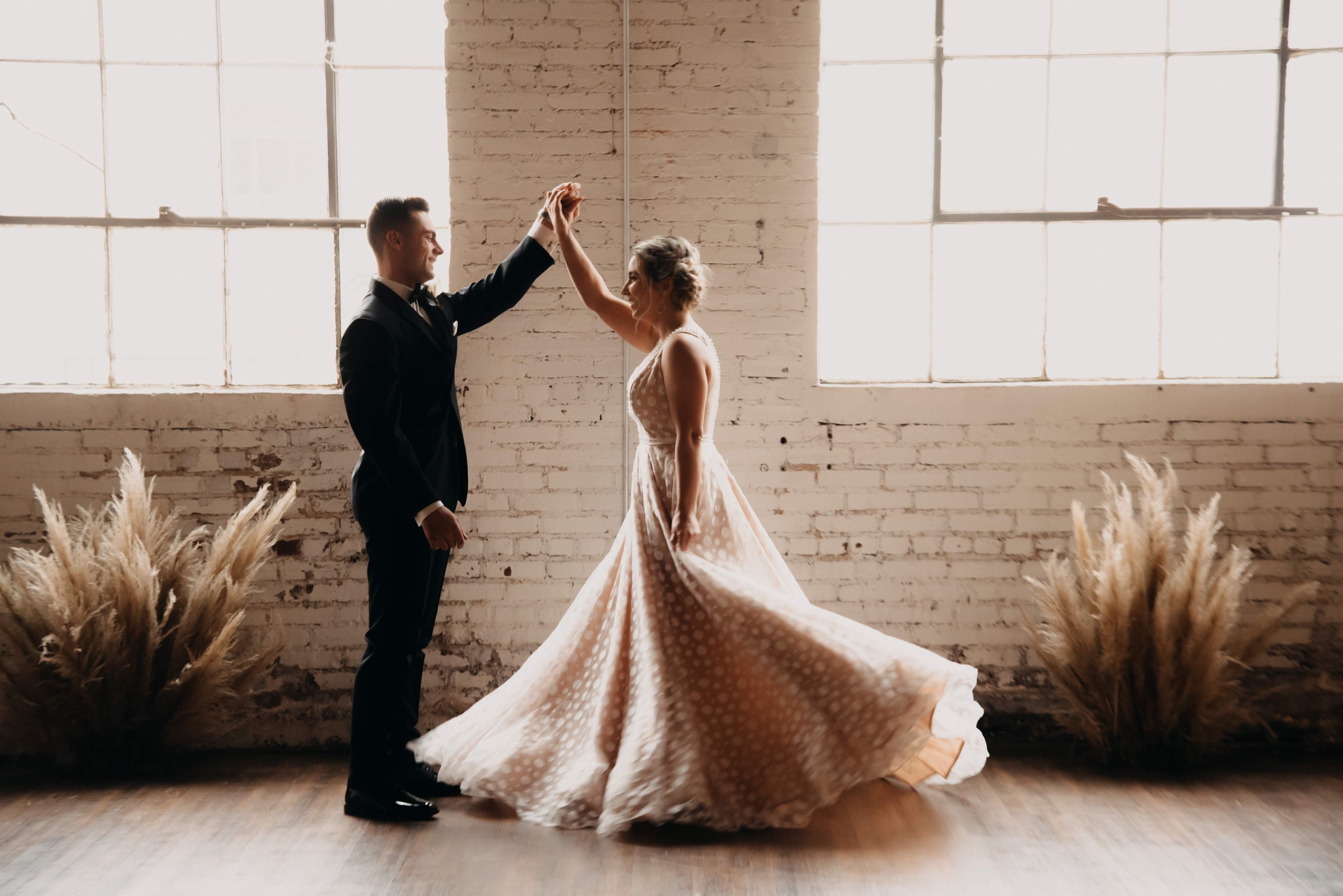 A couple dancing alone in the ballroom in daylight — he turns her beneath his raised hand and her polka-dot gown sweeps the floor, against whitewashed brick, tall factory windows and pampas grass.