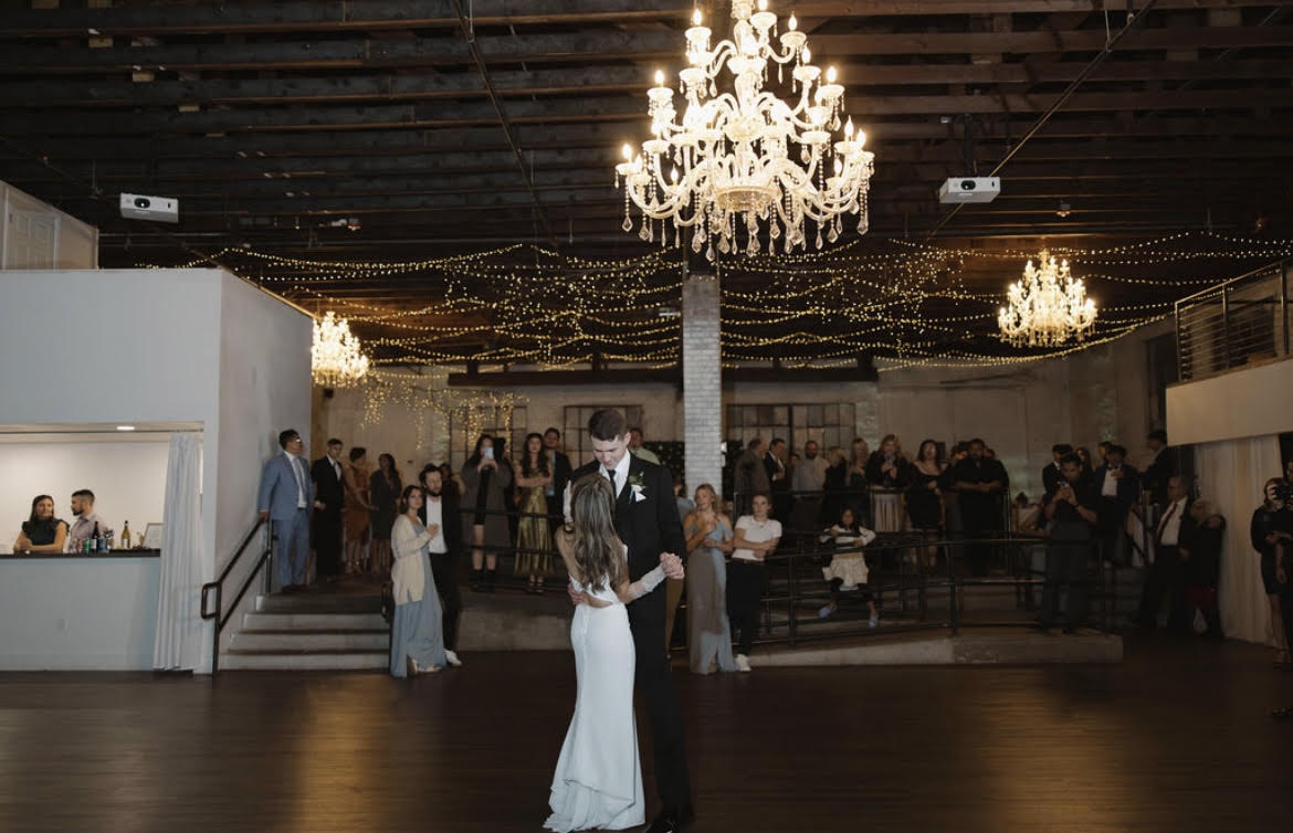 A couple's first dance on the main ballroom floor, three glass chandeliers and fairy lights strung overhead through the original wood trusses while guests watch from the raised perimeter.