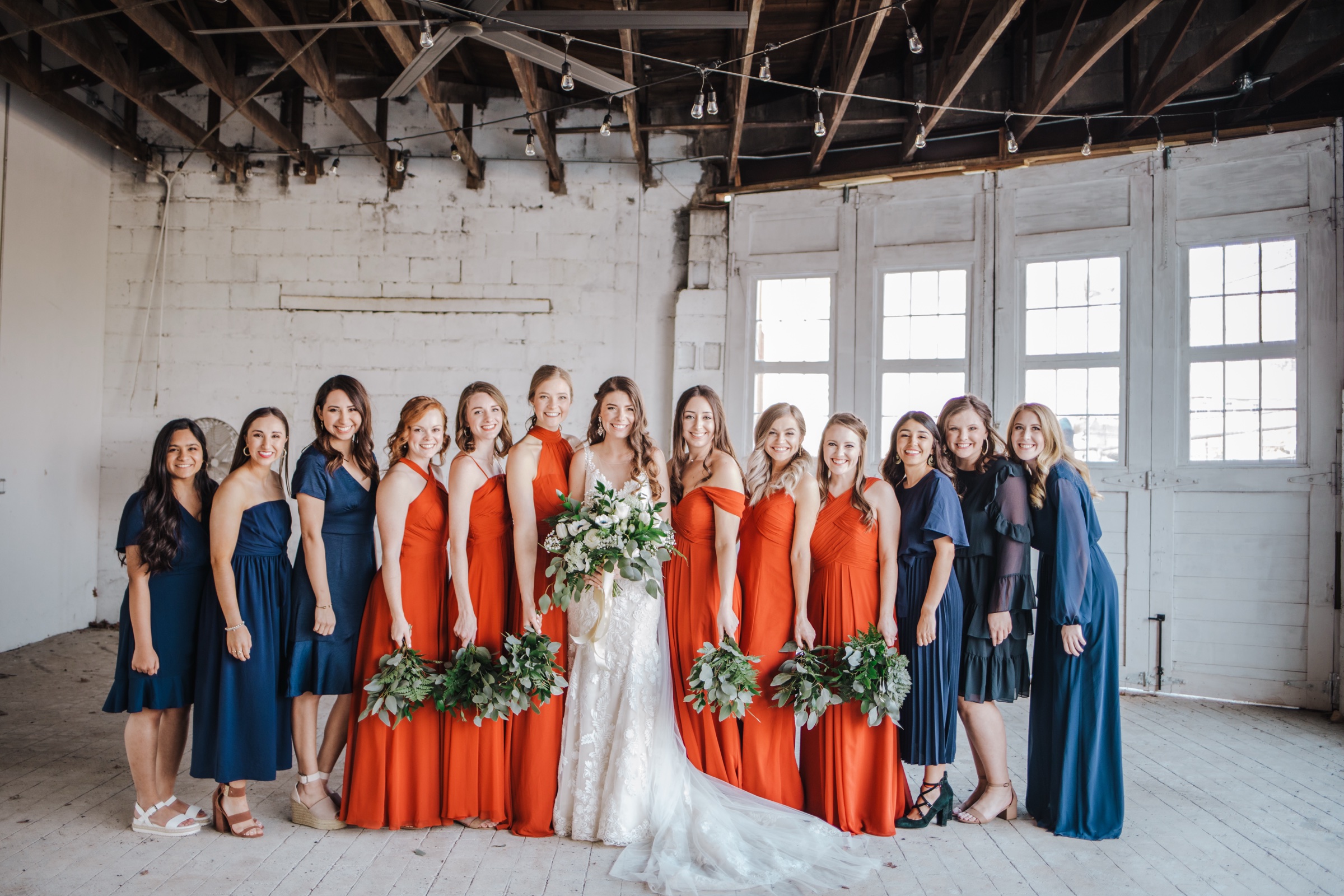 A full bridal party lined up across the ballroom in burnt orange and navy, whitewashed brick and the wood trusses behind them.