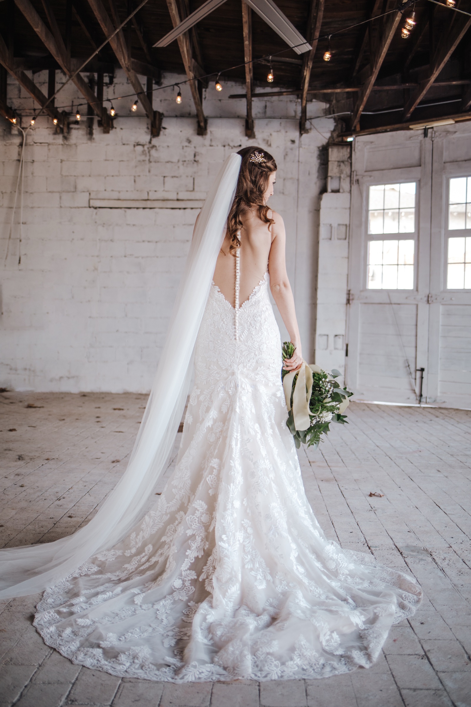 A bride photographed from behind, her lace train and cathedral veil spread across the ballroom floor, whitewashed brick and the tall white doors beyond.