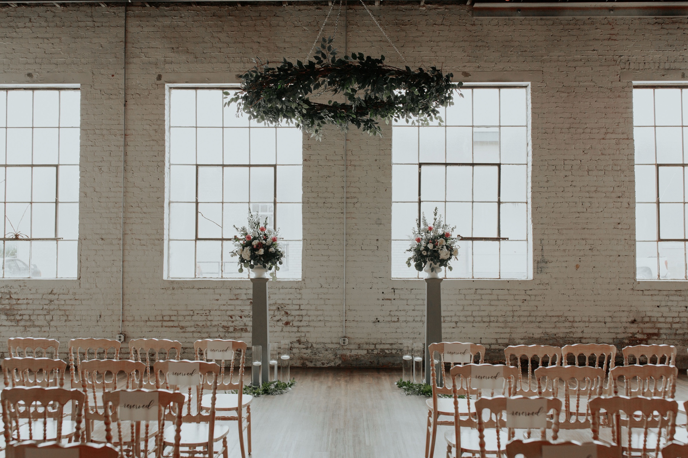 The ballroom set for a ceremony before guests arrive — chiavari chairs either side of the aisle, two raised floral urns, and a ring of greenery hung between the tall factory windows.