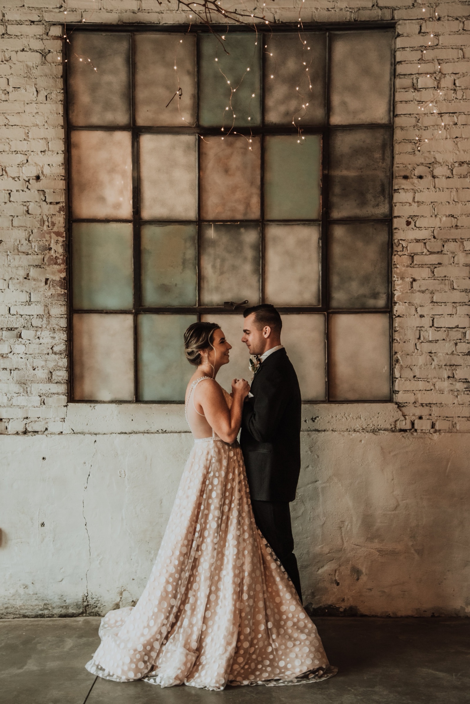 A couple facing each other in front of the venue’s antique multi-pane window, fairy lights strung above and exposed red brick either side.