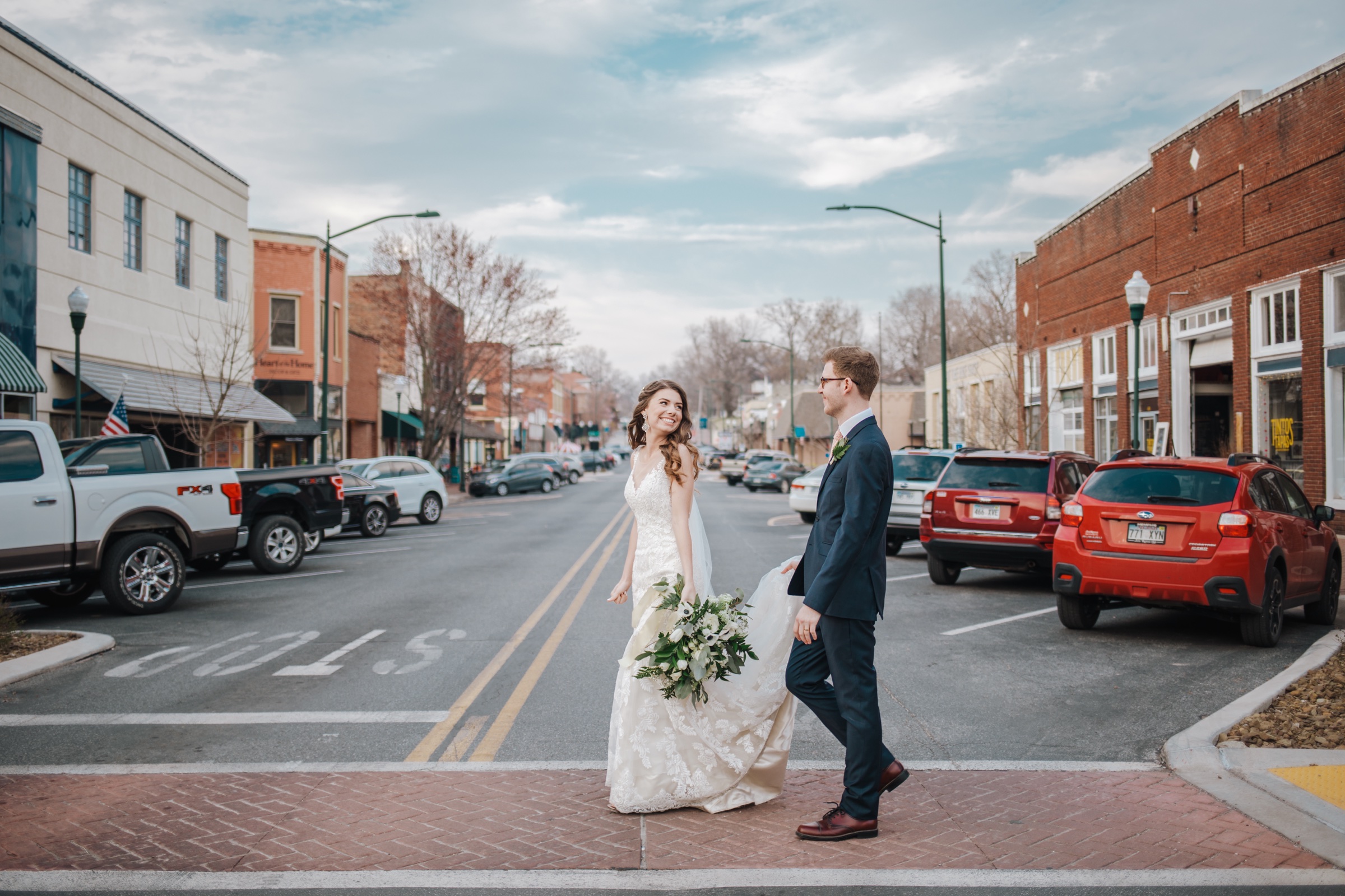 A bride and groom crossing Main Street in downtown Siloam Springs, the brick storefronts of the block running away behind them.