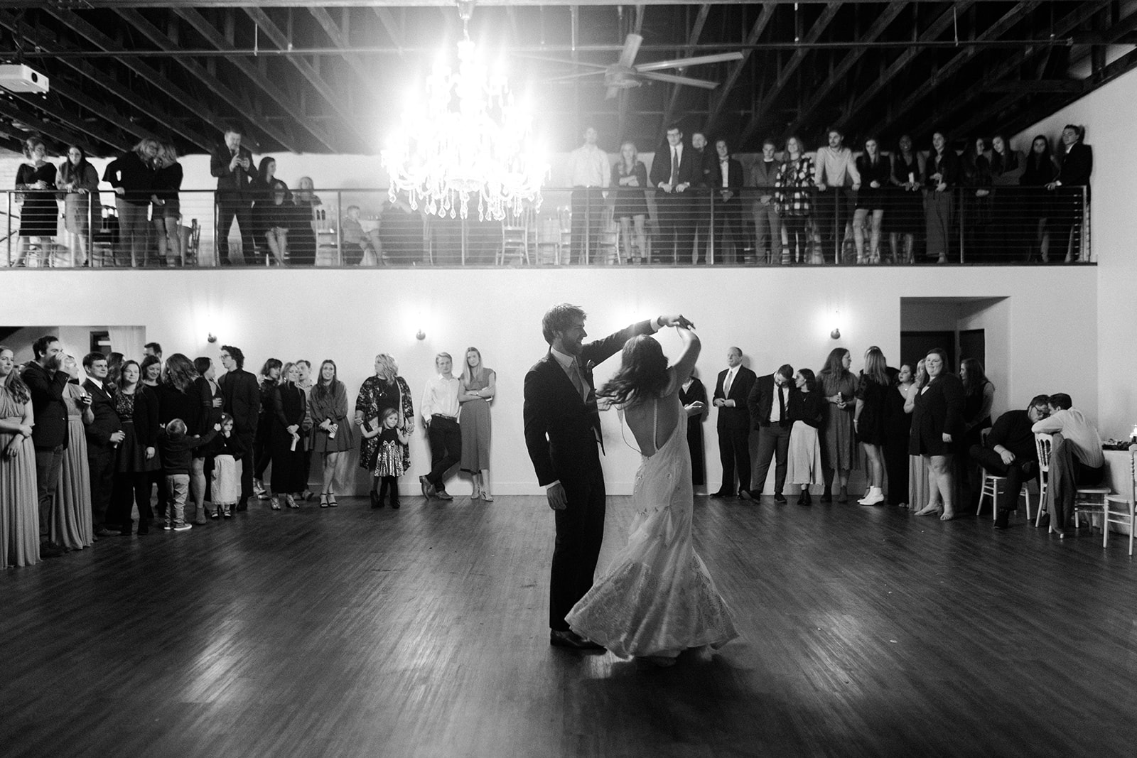 A first dance in the evening, the couple alone on the ballroom floor while guests watch from the floor and the mezzanine above, the chandelier lit overhead. Photographed in black and white.