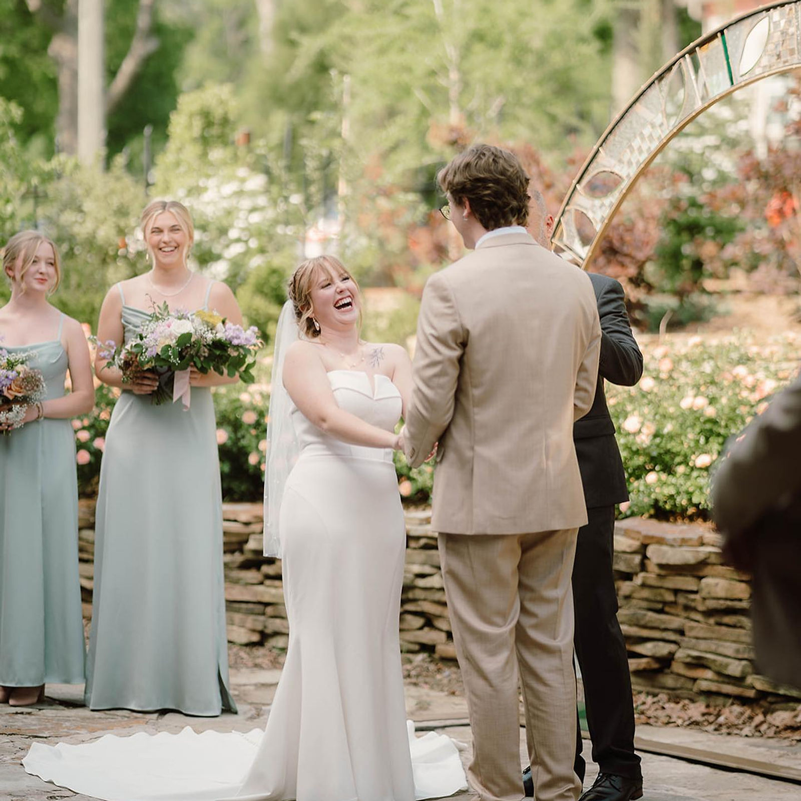 An outdoor garden ceremony — the couple exchanging vows beneath a mosaic arch, roses and a stacked-stone wall behind.