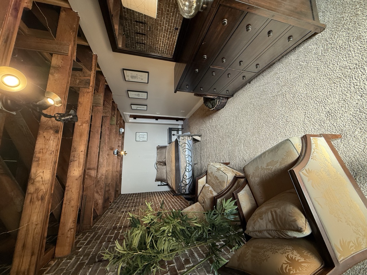A bedroom along a brick wall, the bed at the far end past a long wooden dresser and a potted plant.