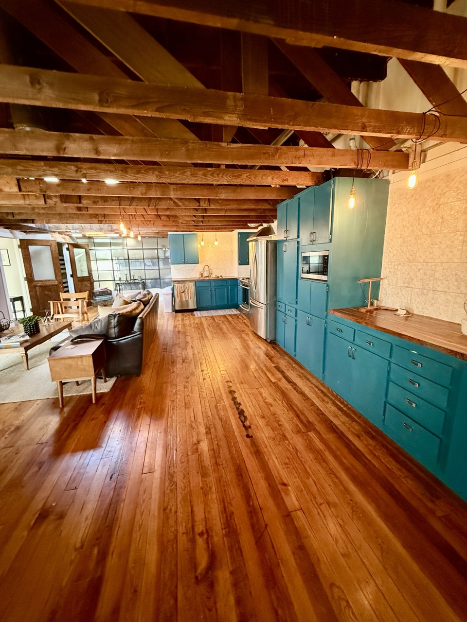 The loft kitchen in teal cabinetry beneath heavy exposed roof trusses, honey-coloured wood floors running the length of the room.