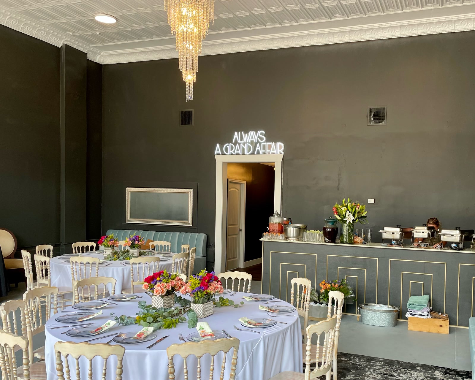 The lobby dressed for a shower: two round tables in white linen with bright flowers, under a crystal chandelier and a pressed-tin ceiling, with the charcoal-and-gold bar laid out as a buffet behind them.