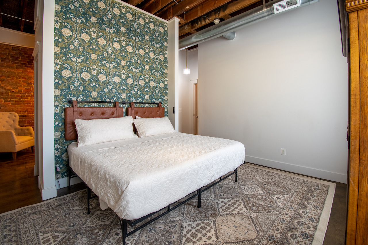 A bedroom with a botanical wallpaper feature wall behind the bed, an antique wooden armoire beside it, and a patterned rug underfoot.