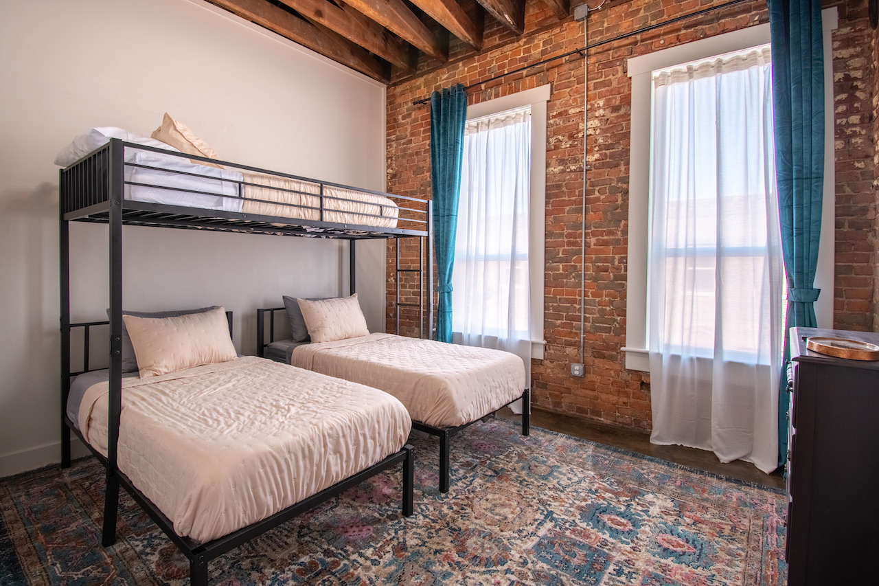 The bunk room: a raised loft bunk above two twin beds against bare brick, teal curtains at the tall windows.