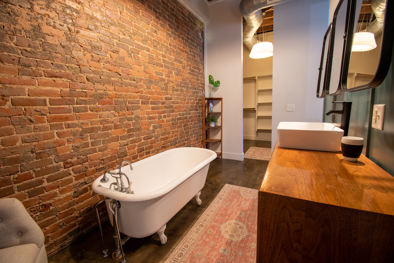 A bathroom with a freestanding clawfoot tub against a full wall of bare brick, and a vessel sink set on a live-edge wood counter.