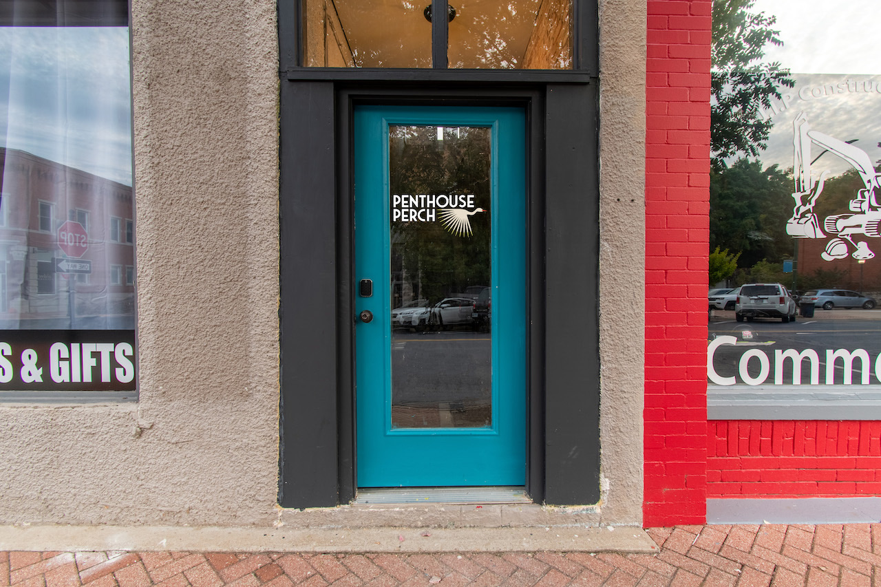 The teal front door to Penthouse Perch, its name etched on the glass, between downtown storefronts on South Broadway.