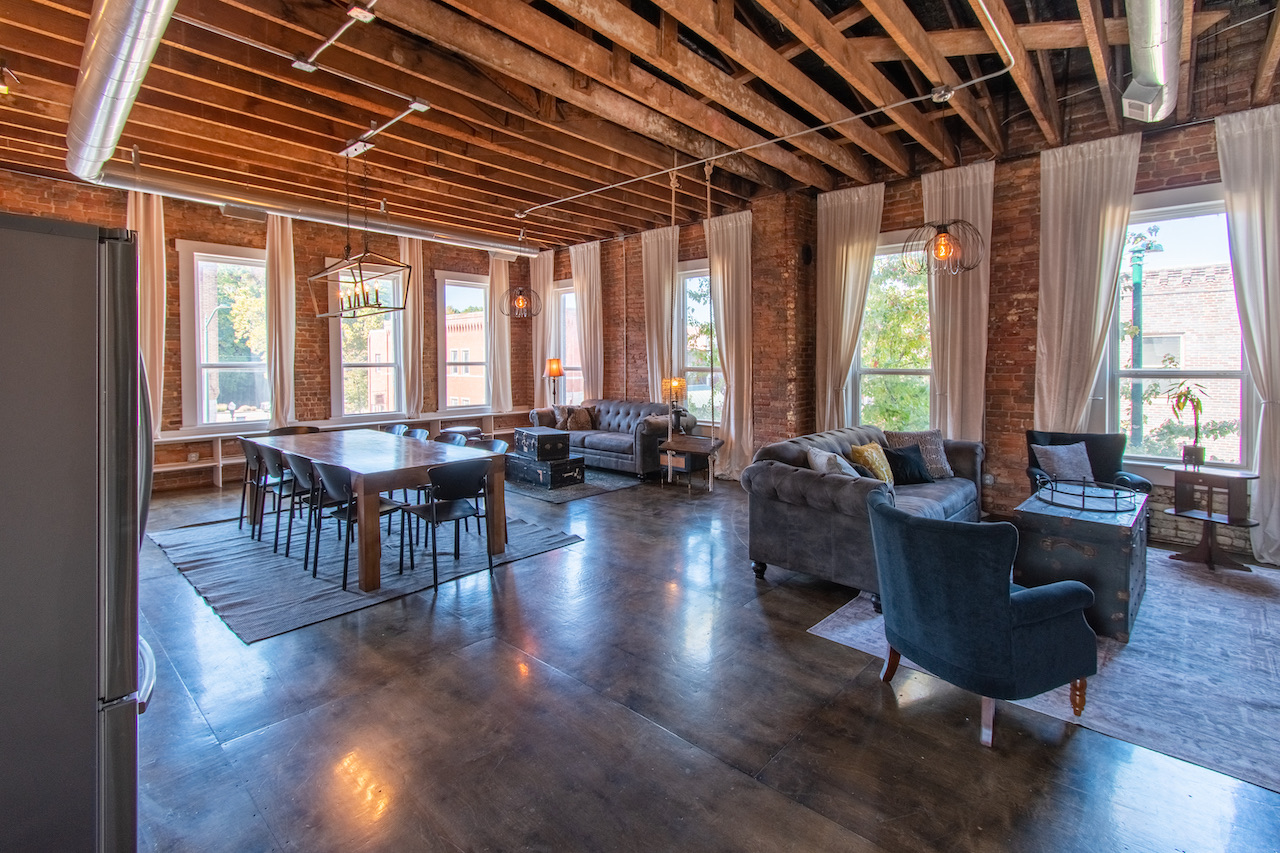 Penthouse Perch's open living room: exposed wood joists, red brick, tall curtained windows over downtown, and tufted grey sofas on polished concrete.