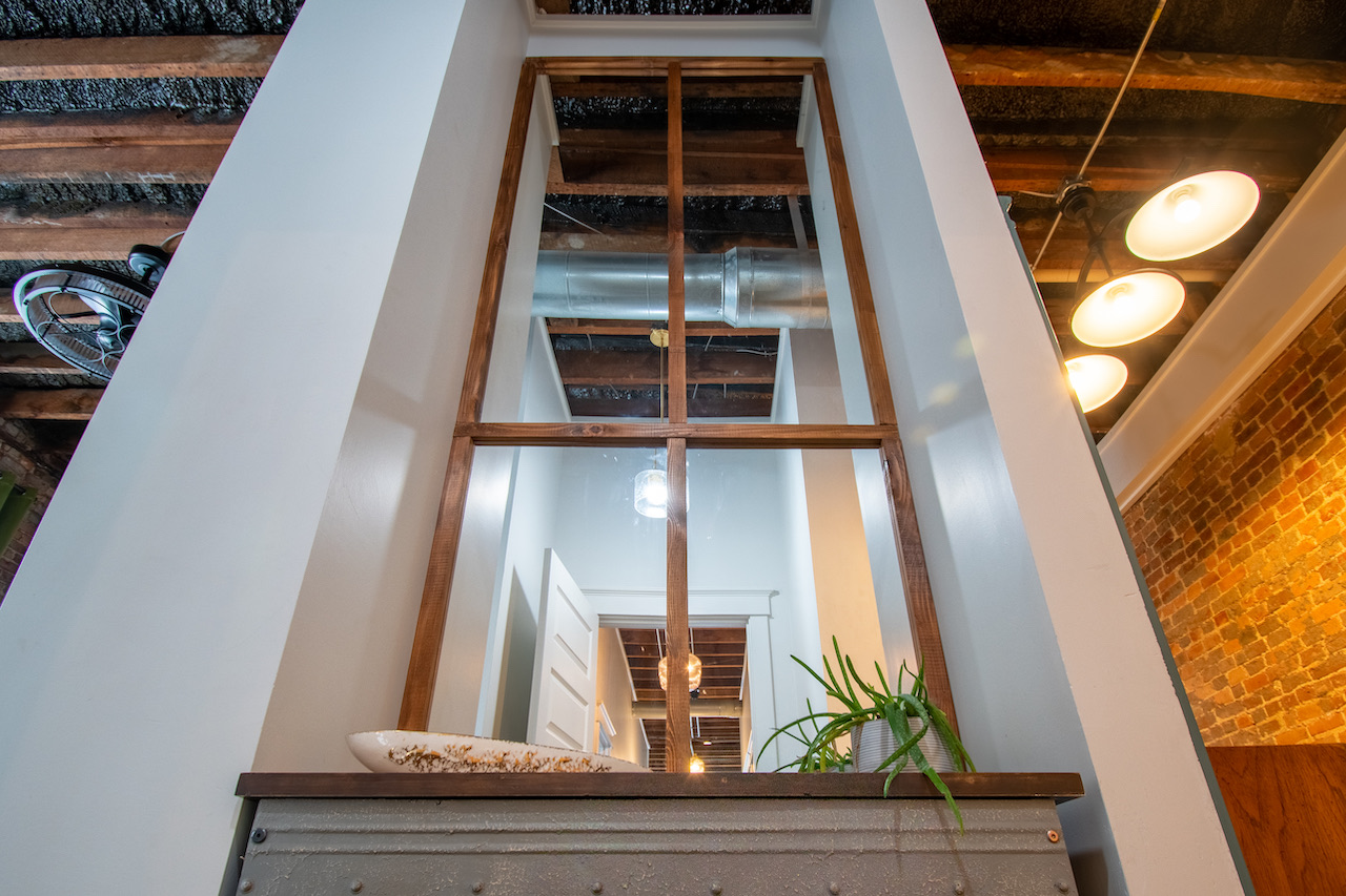 The stairwell, looking up past exposed joists and pendant bulbs to a tall multi-pane window.