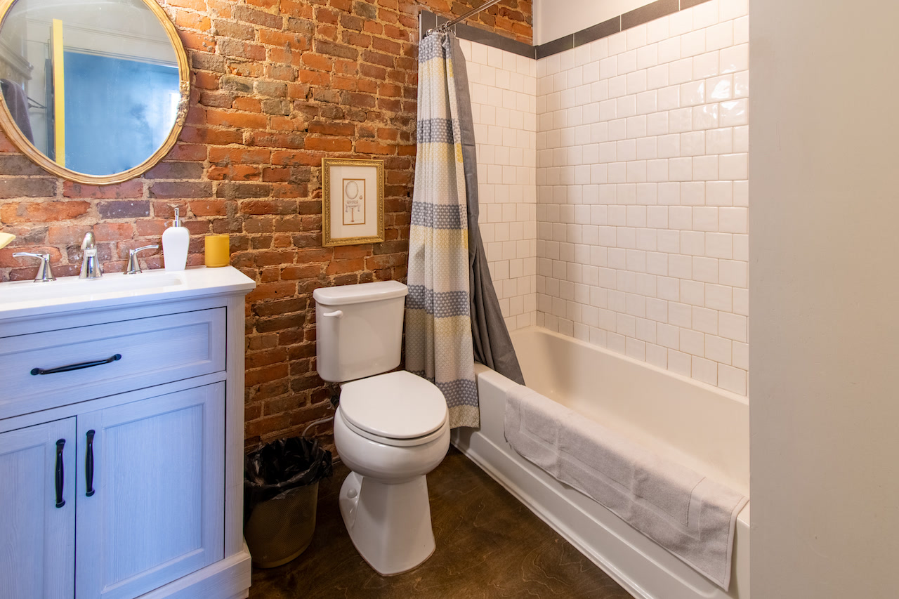 The second bathroom: a pale blue vanity and round gold-framed mirror against exposed red brick, next to a white subway-tiled tub and shower.