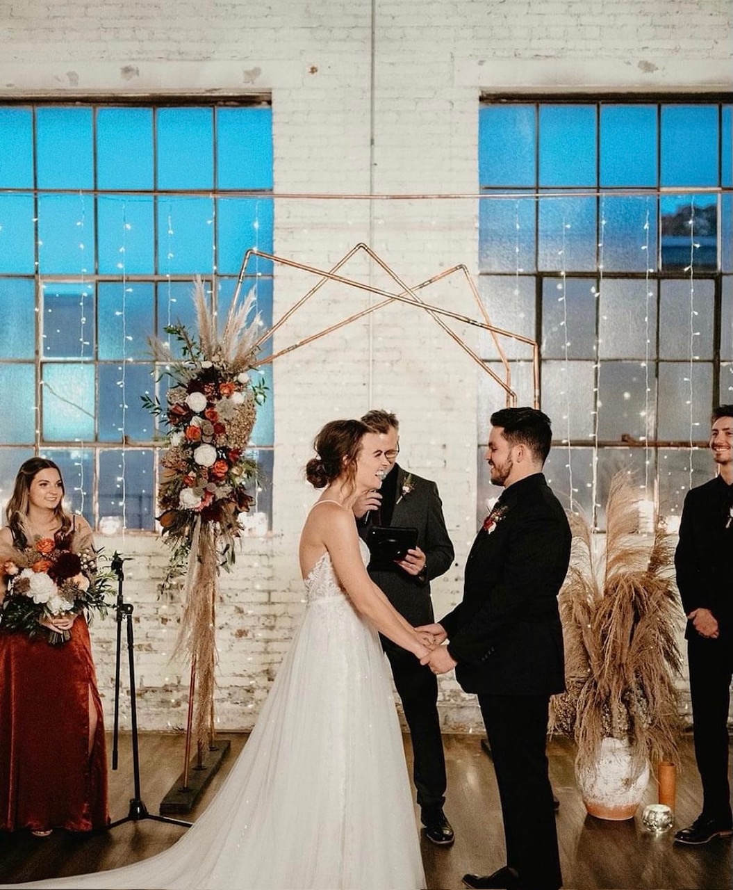The metal triad arbor: three slim gold uprights behind a couple at their ceremony, dressed with pampas and dried florals.