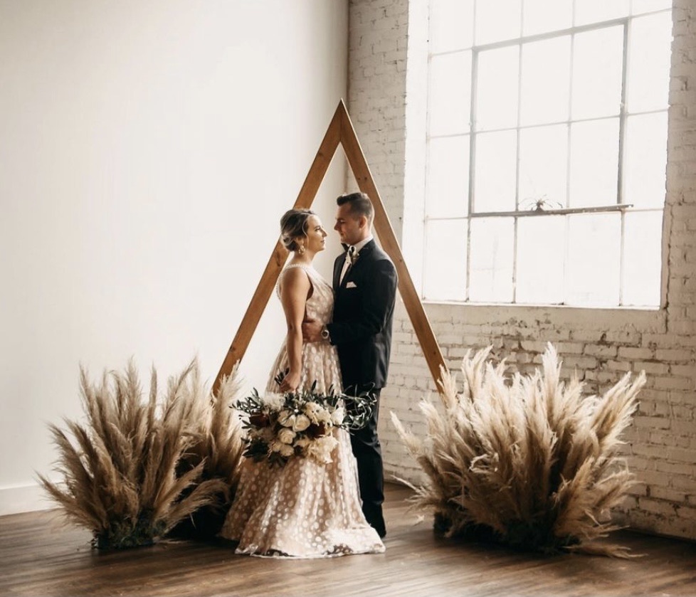 The triangle arbor: a tall gold triangular frame against whitewashed brick, flanked by large pampas grass arrangements.
