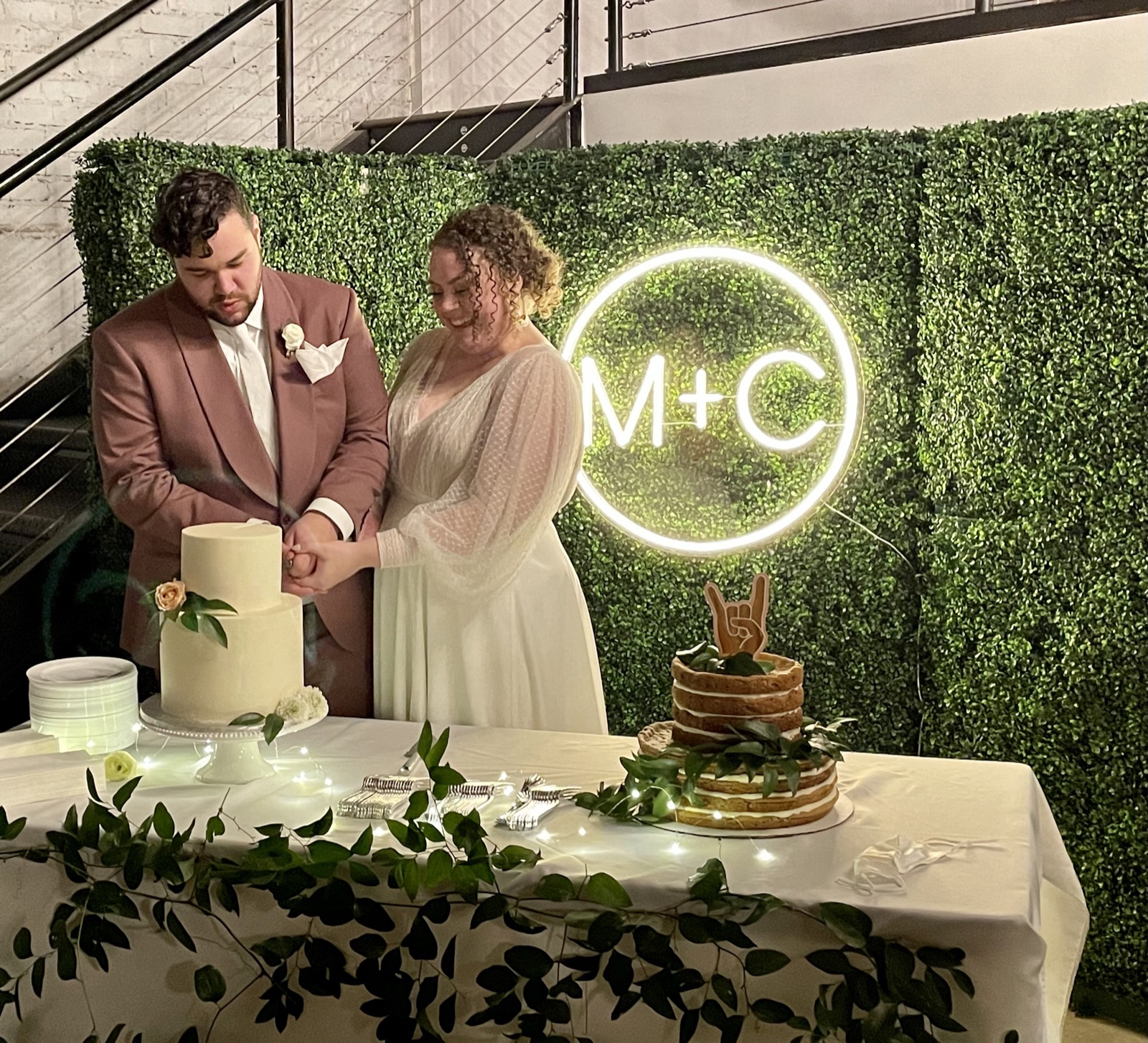 A couple cutting their cake in front of a green hedge wall hung with a neon monogram sign, the cake table trimmed with greenery.