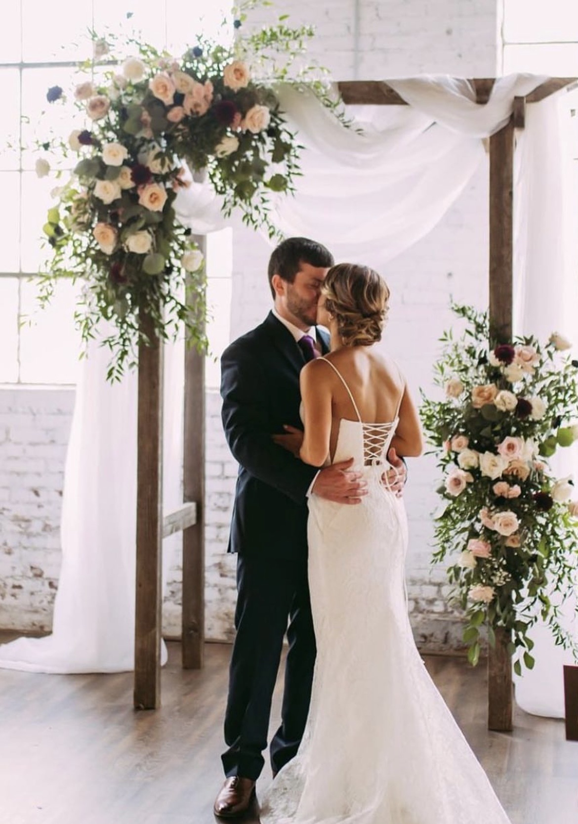 A couple embracing beneath a wooden arbor dressed with white drapery and blush floral clusters at its corners.