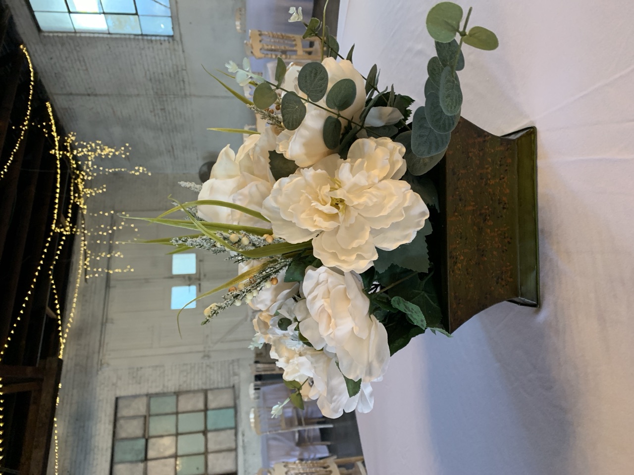 A white rose and greenery arrangement in a dark metal footed planter, on a round table under the ballroom's fairy lights.