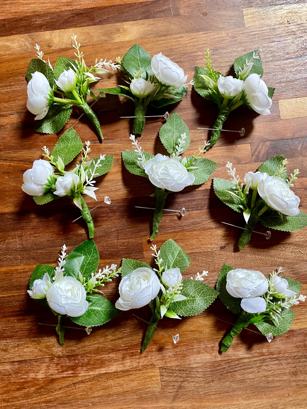 Nine matching white rose boutonnieres with green leaves and pins, laid out in rows on a wooden table.
