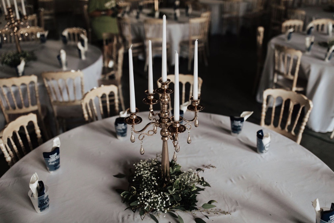 A tall branched candelabra dressed with greenery, standing on a linen-covered round table set with glassware.
