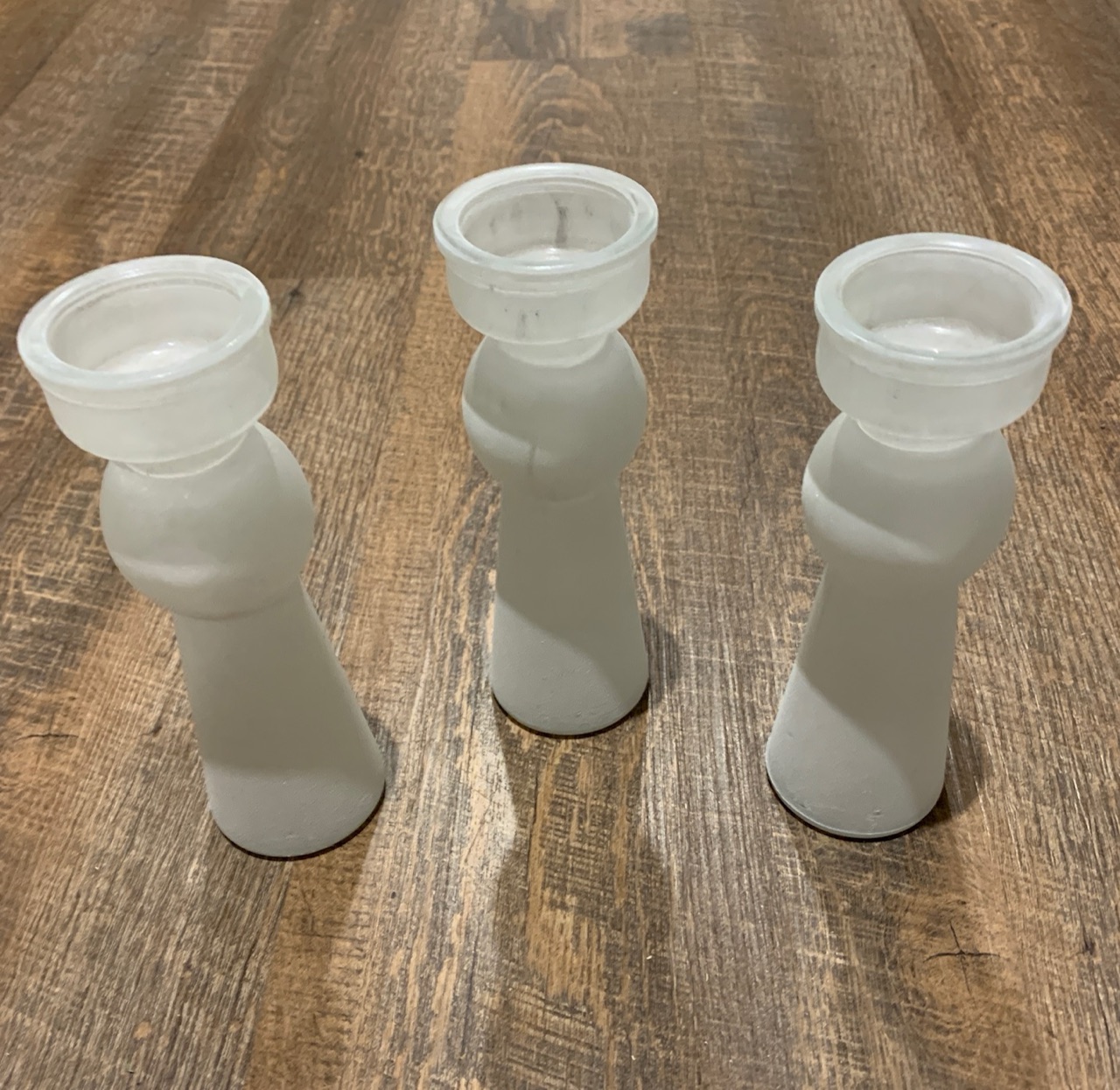Three frosted white glass candle holders of the same shape, stood in a row on a wood floor.