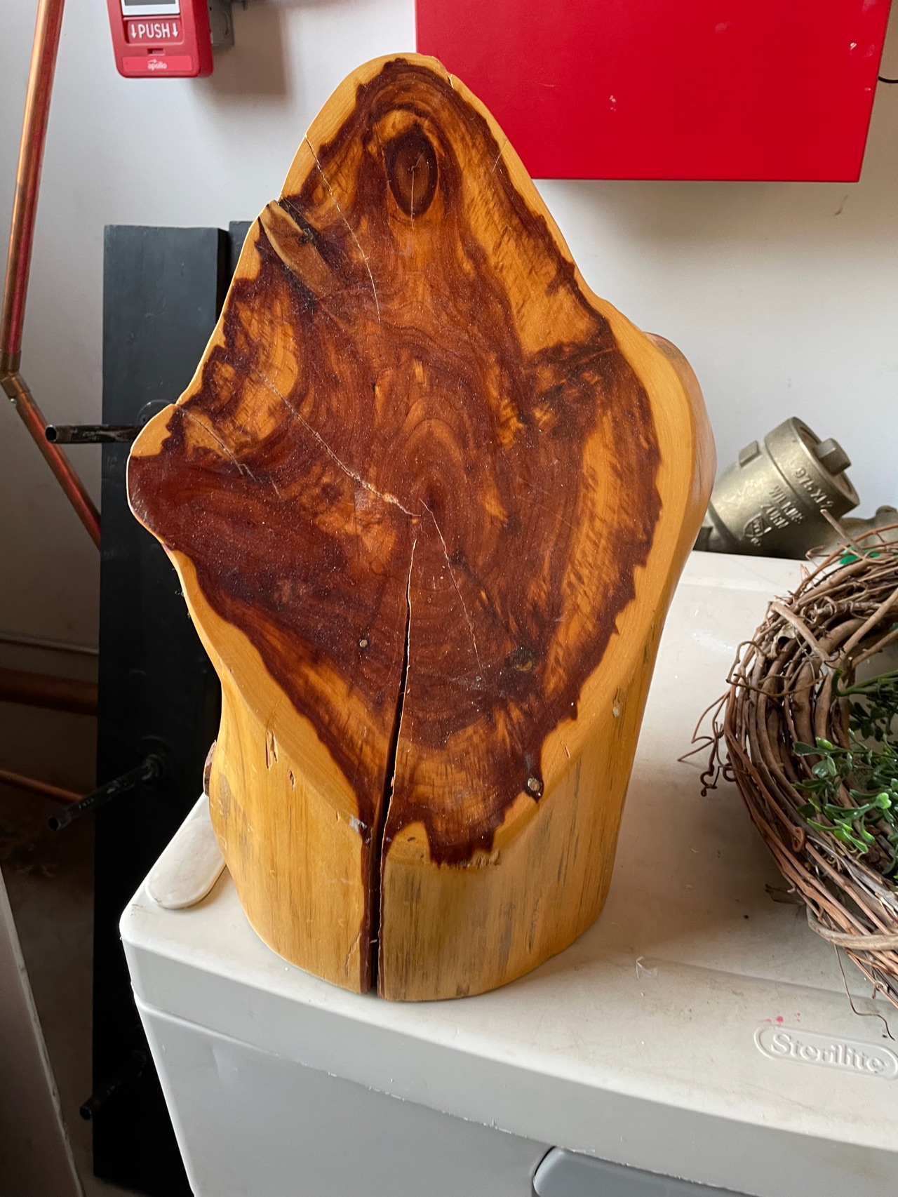 A polished live-edge cedar slab with deep red grain, stood upright on a table with a grapevine wreath behind it.