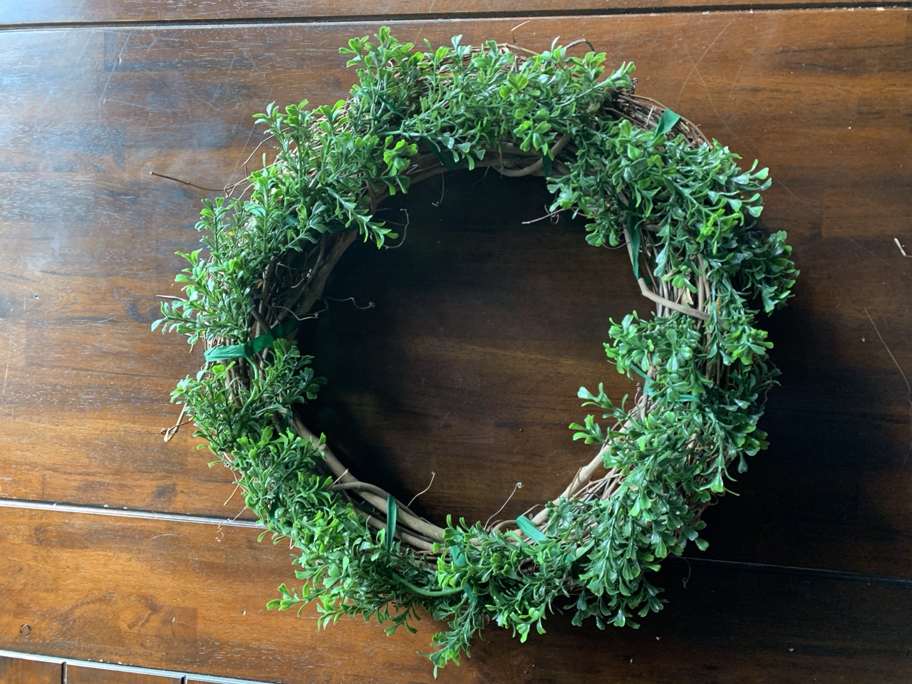 A bright green boxwood wreath on a grapevine base, laid on a dark wood table.
