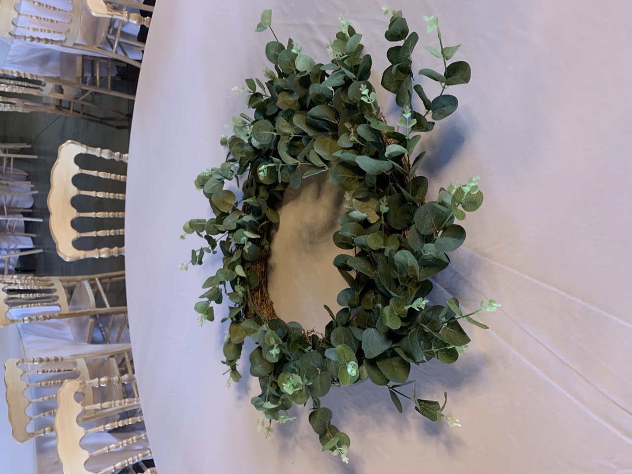 A round eucalyptus wreath laid flat as a centerpiece on a white linen round table.