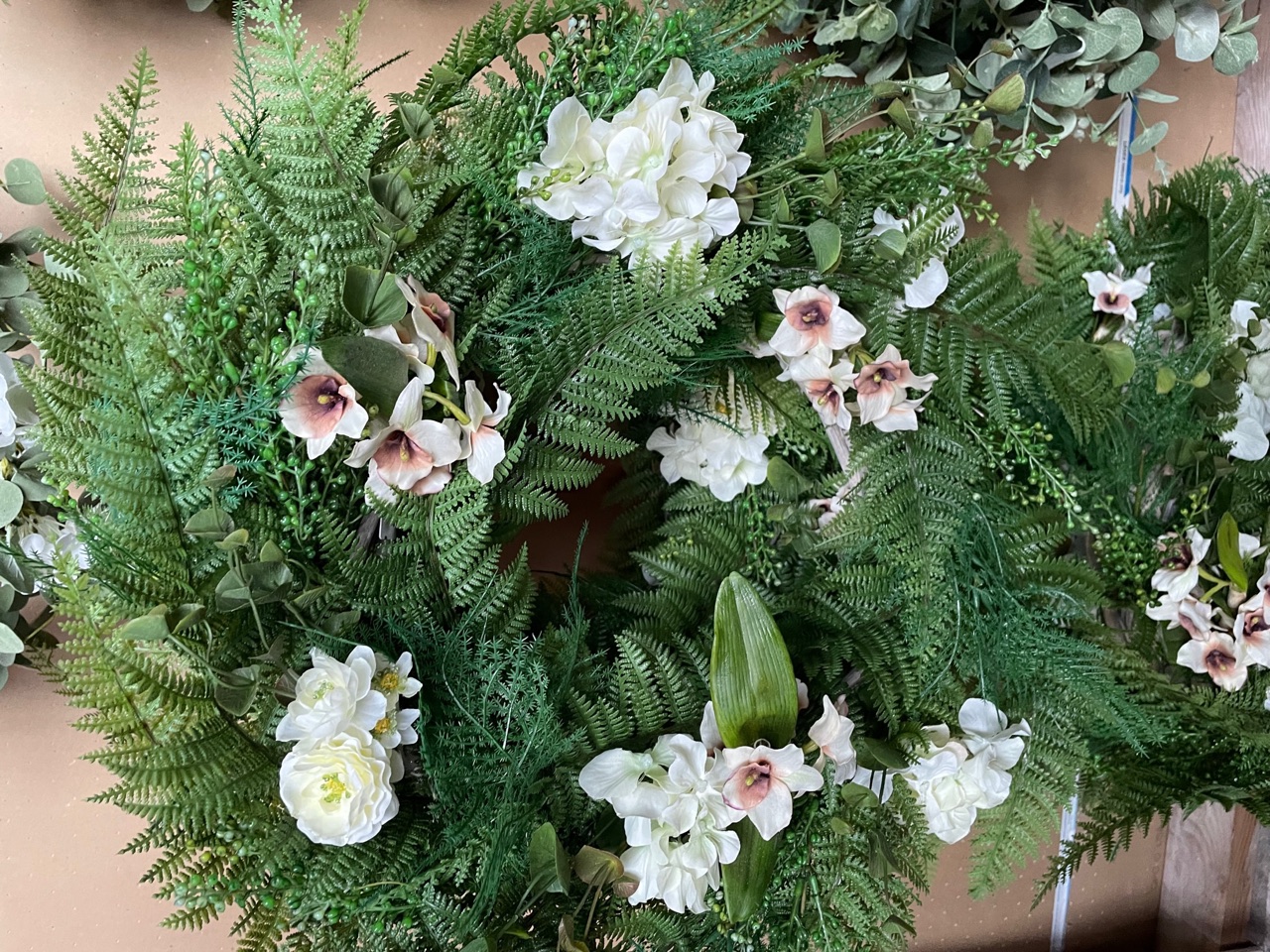 A full fern wreath studded with white and blush silk blooms, against a brick wall.