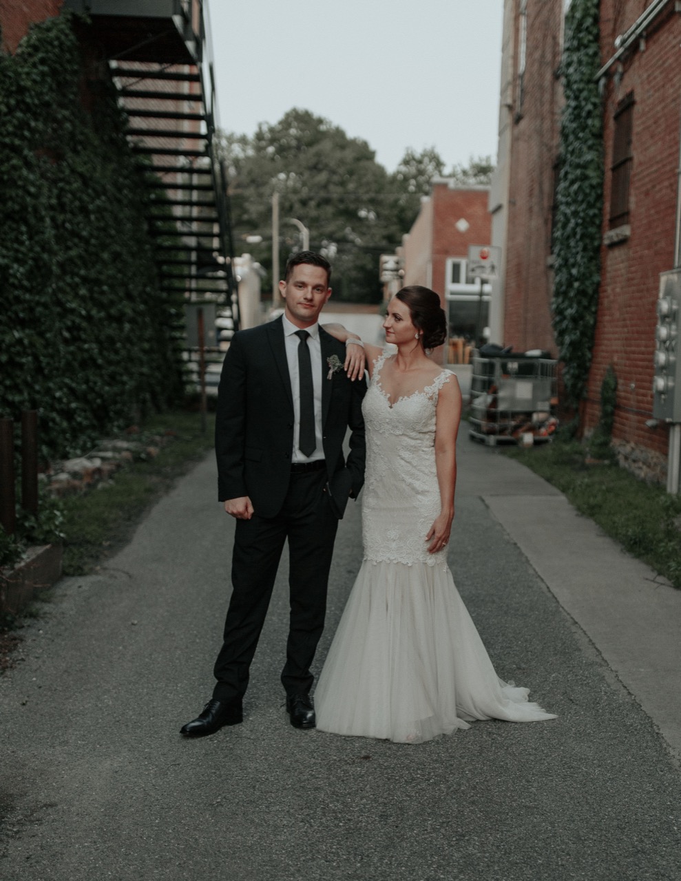 A couple standing together in the alley, ivy climbing the brick and an iron fire escape overhead.