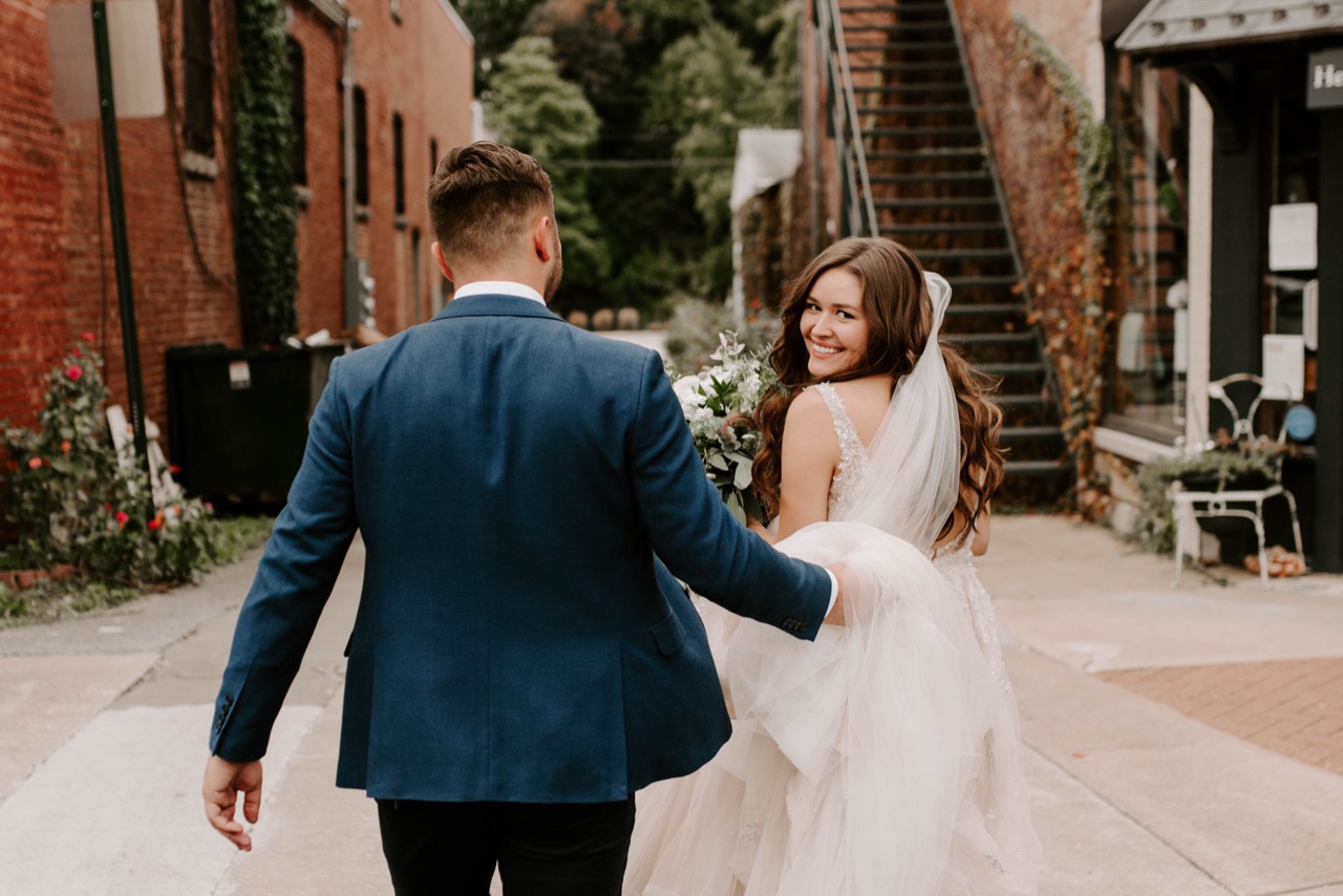 The bride glancing back over her shoulder as the couple walk the ivy-covered alley beside the building.