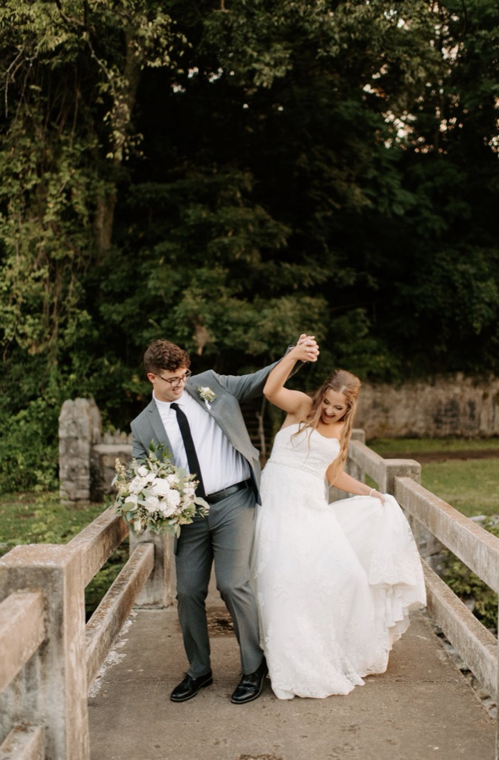 The groom twirling the bride on the stone bridge, summer greenery closing in on both sides.