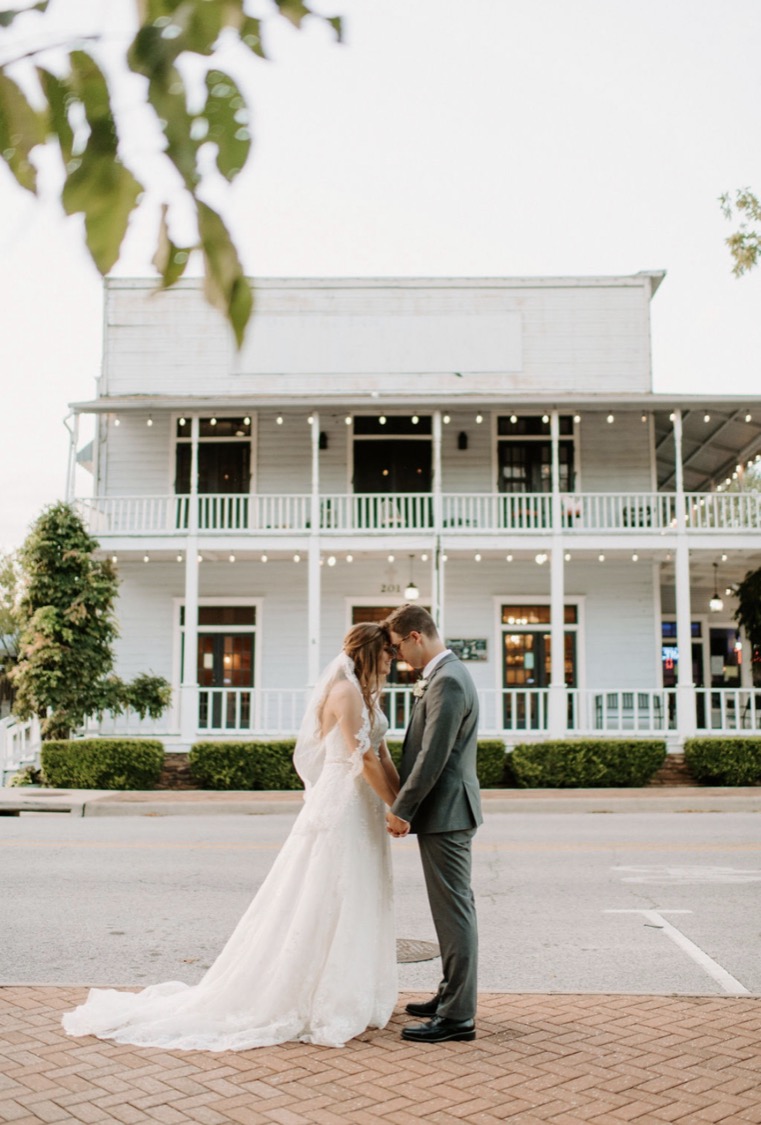 A couple forehead to forehead in the street, a white two-storey porch strung with lights behind them.