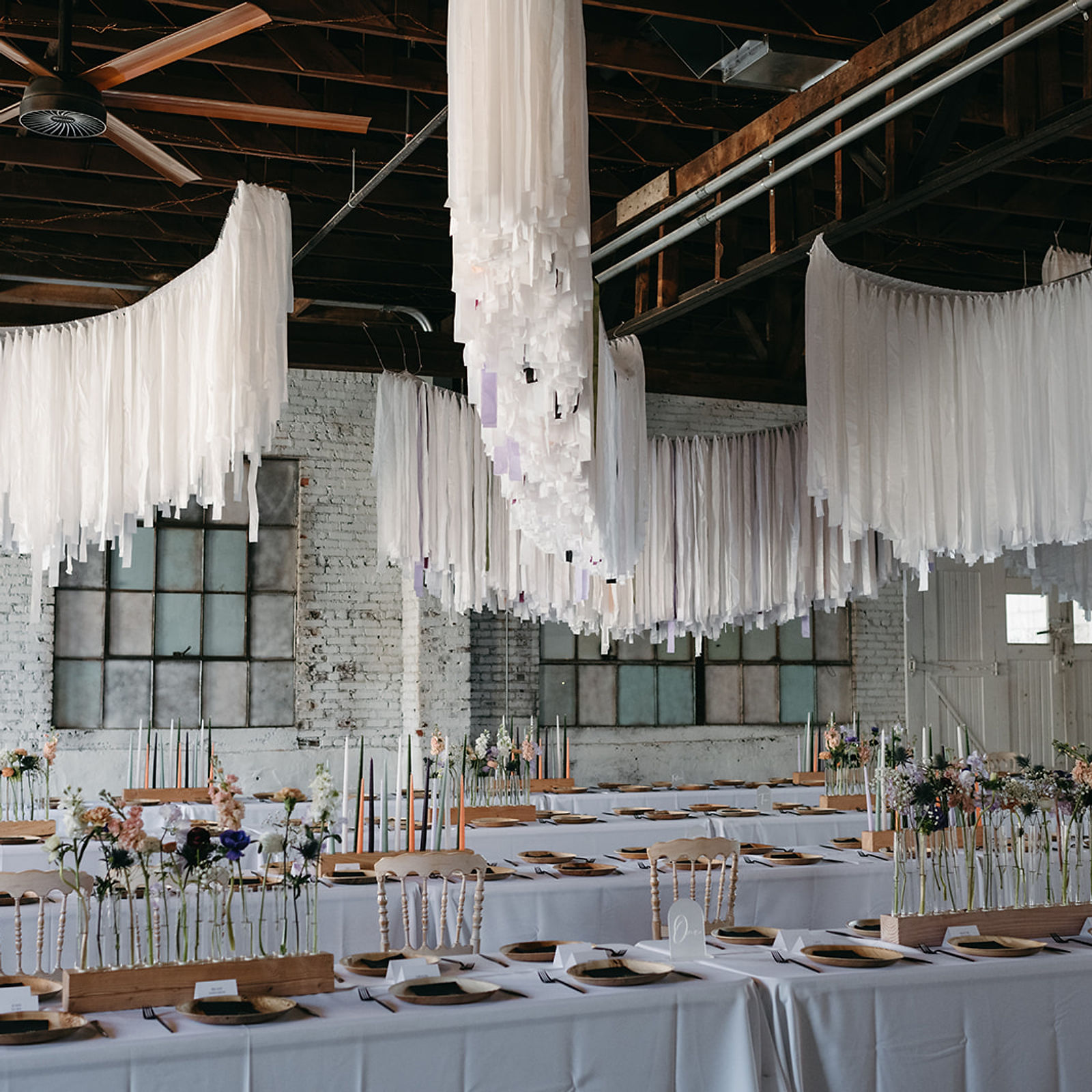 Long reception tables set beneath a hanging fabric installation, whitewashed brick behind.