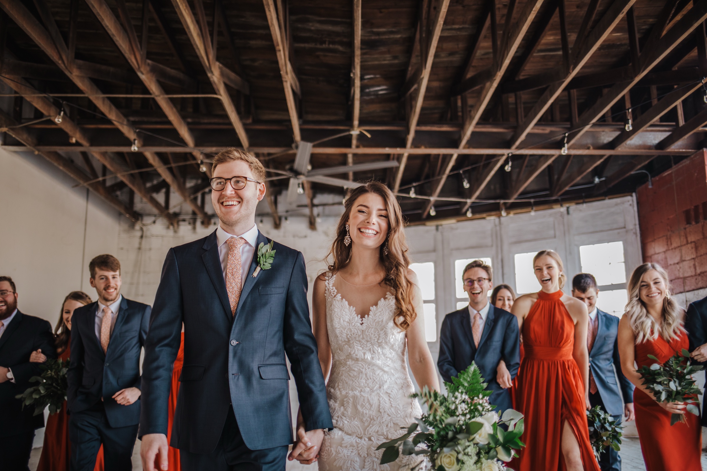 A couple walking back down the aisle hand in hand after the ceremony, their wedding party following in navy and burnt orange, beneath the ballroom’s original wood trusses.