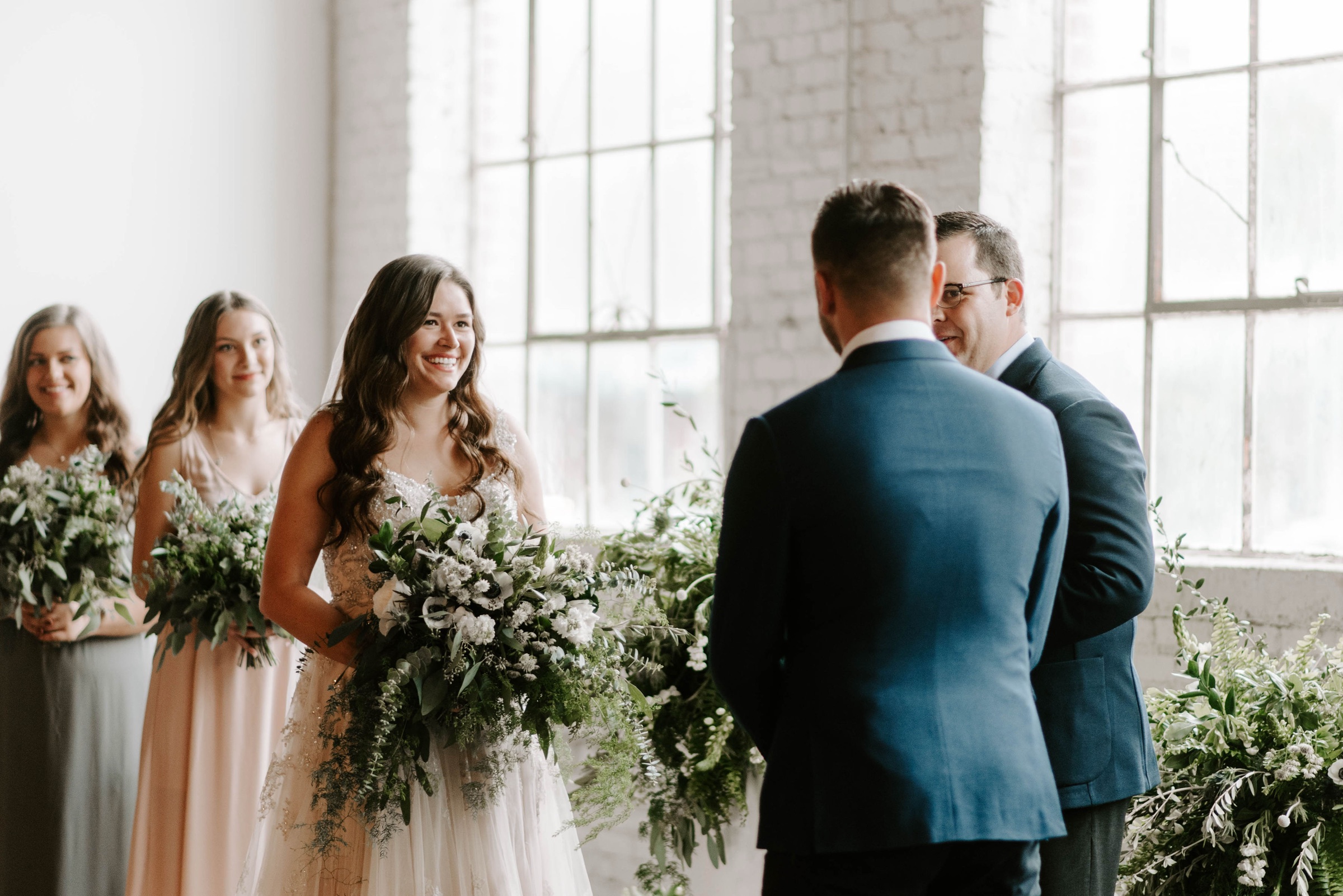 A couple exchanging vows in front of their officiant, bridesmaids holding bouquets to one side, backlit by the ballroom’s tall windows.